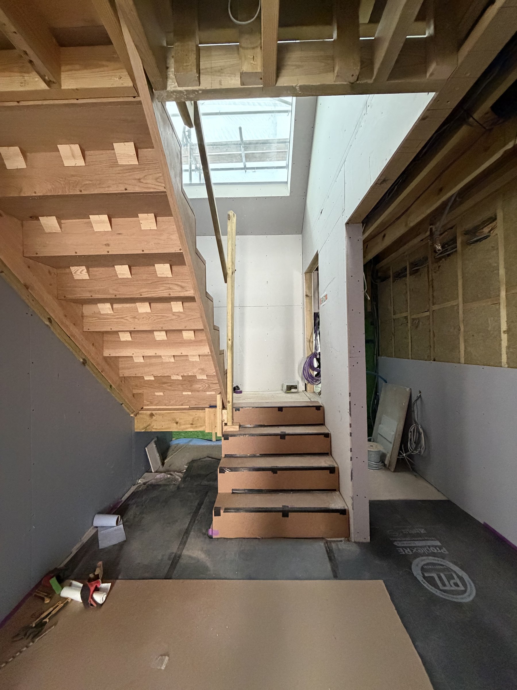 Solid oak staircase with its underside exposed rising past a rooflit stairwell, treads under protective board