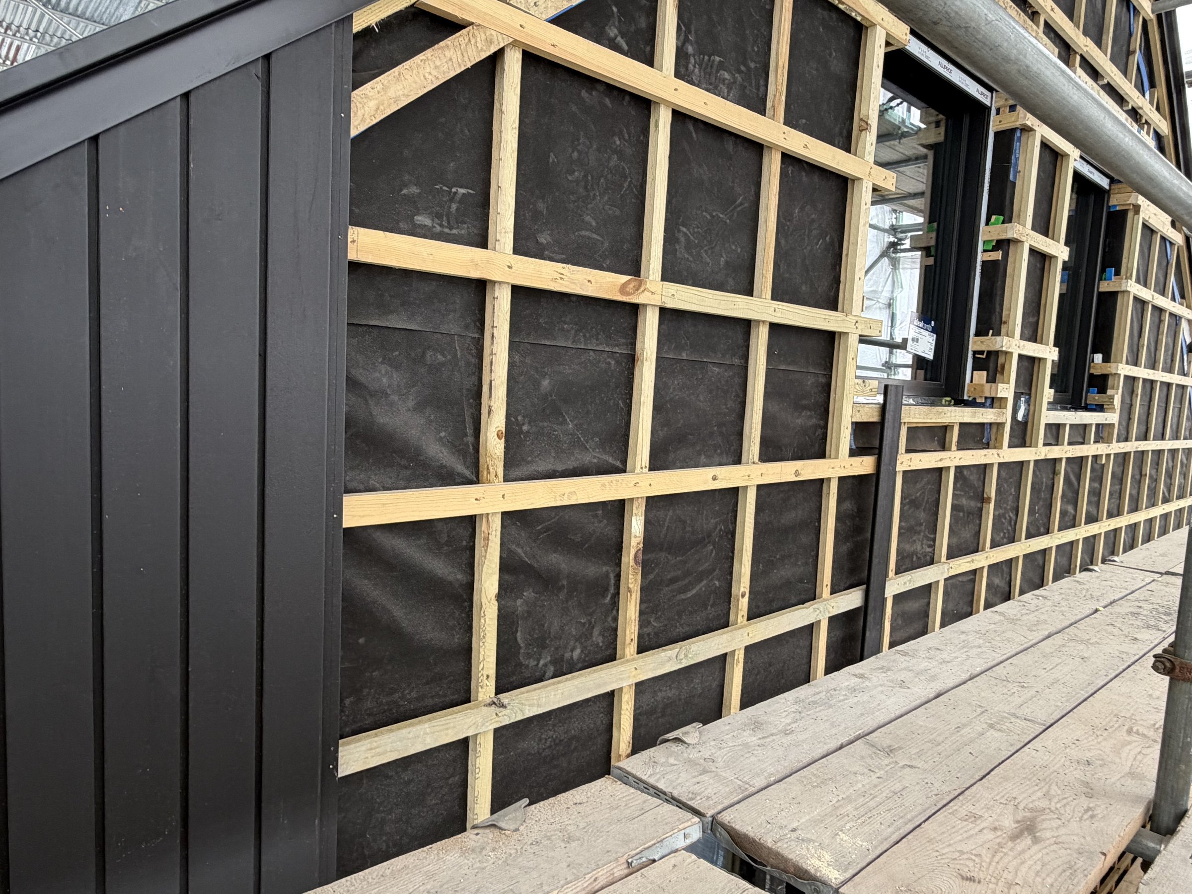 Black vertical cladding boards fixed beside a grid of timber battens over black breather membrane, windows beyond