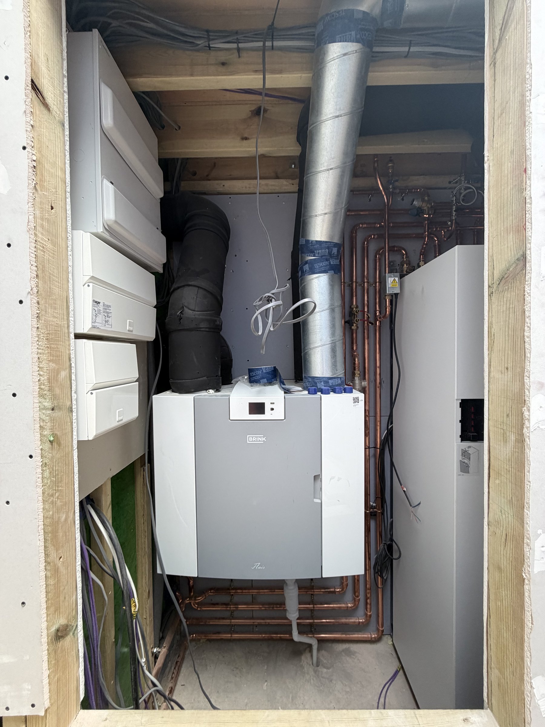 Heat recovery ventilation unit in the plant cupboard with taped ducts above it and copper heating pipework beside it