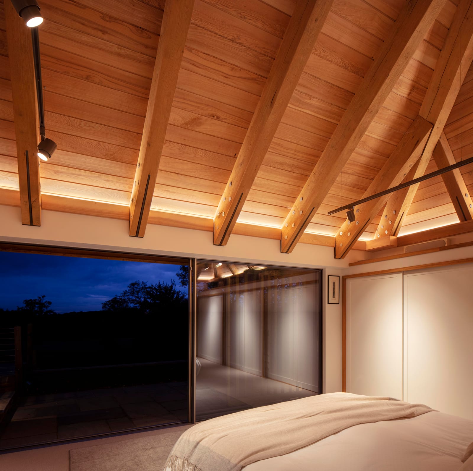 Bedroom under an exposed timber roof with sliding glass to the terrace at dusk