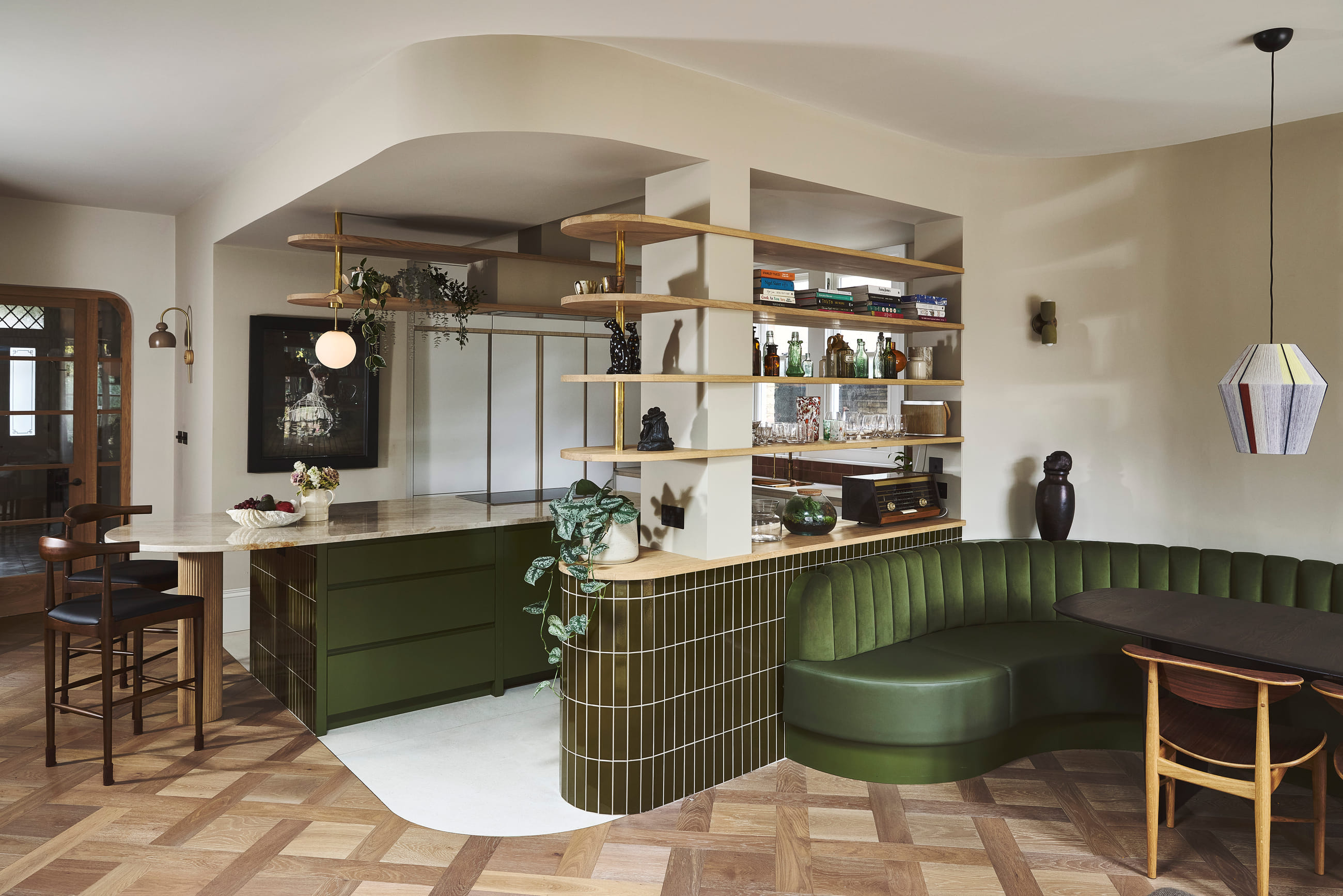 Open kitchen with green cabinetry, a glazed tile island, and a curved banquette under oak and brass shelving