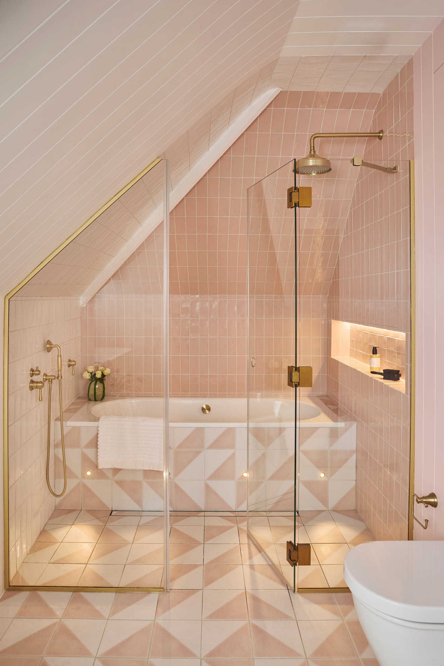 Bathroom under the eaves, tiled in blush pink with a shower over the bath