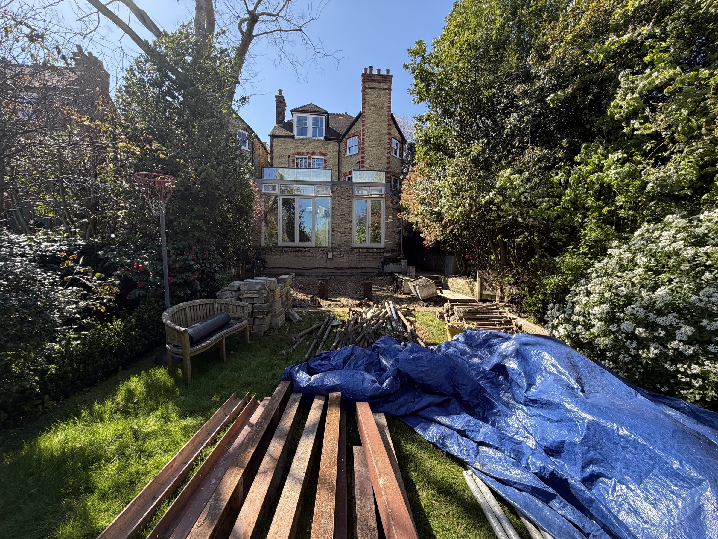 Structural steel sections laid out on the rear lawn, the deck removed and its brick sub-structure exposed at the house
