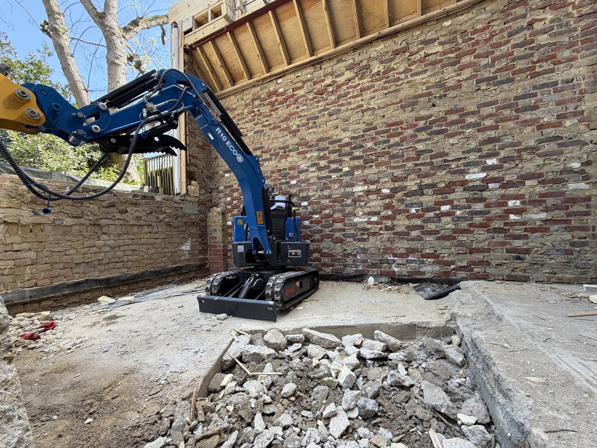 Mini excavator breaking out the old concrete floor slab inside the stripped rear addition