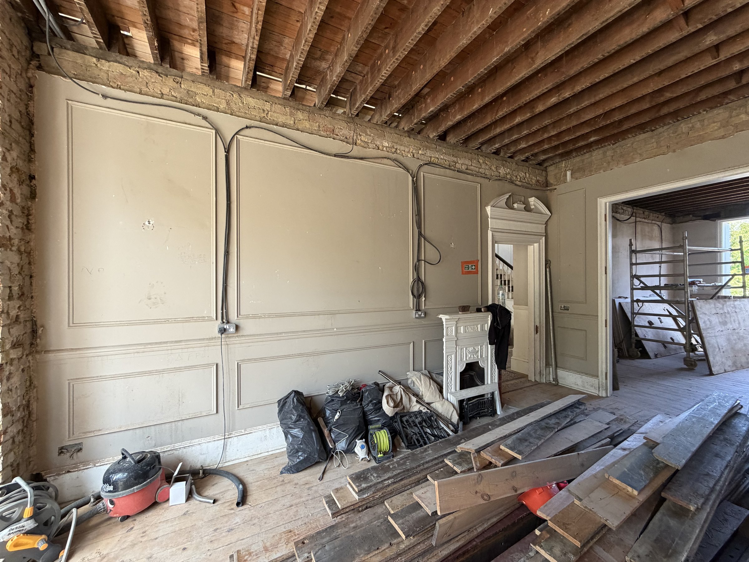 Ground floor reception room mid strip-out, painted panelling retained on one wall, ceiling joists exposed, cast iron fire surround set aside